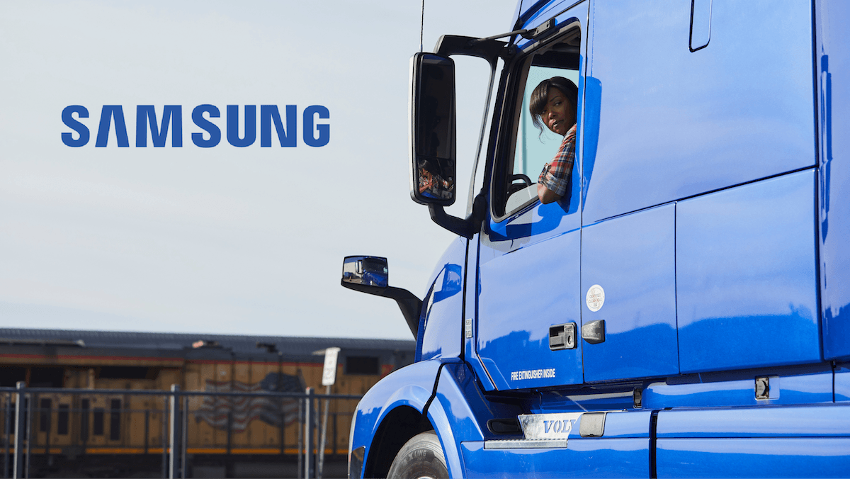 Truck driver looking out the window of a bright blue semi‑truck cab with a freight train in the background.